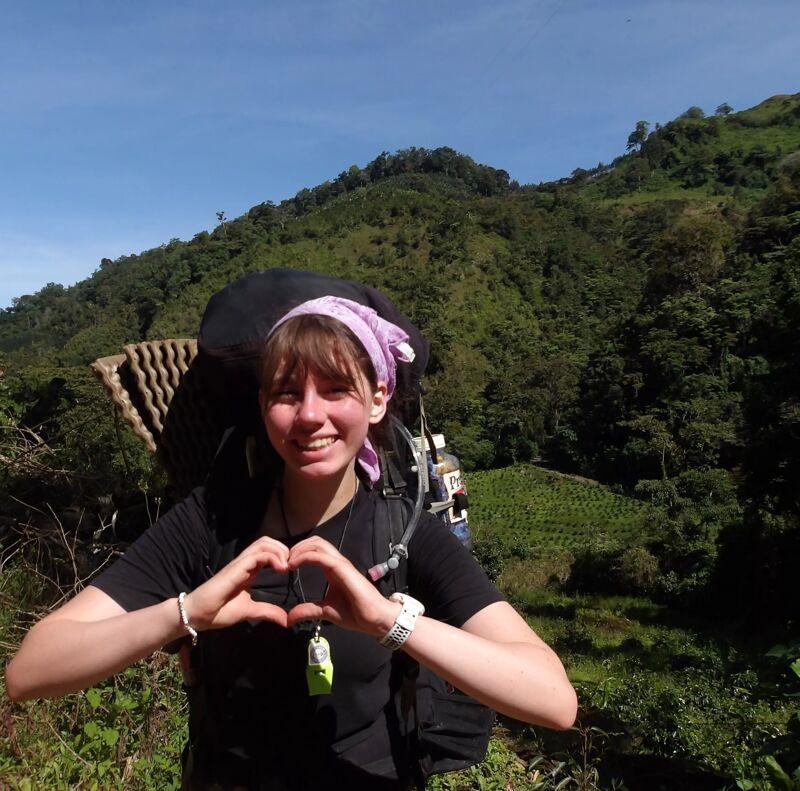 A young woman with a backpack is making a heart shape with her hands. She is wearing a black t-shirt and a patterned headband. Behind her is a lush green hillside with trees and vegetation. The sky is blue and clear. She appears to be hiking or backpacking in a mountainous area.